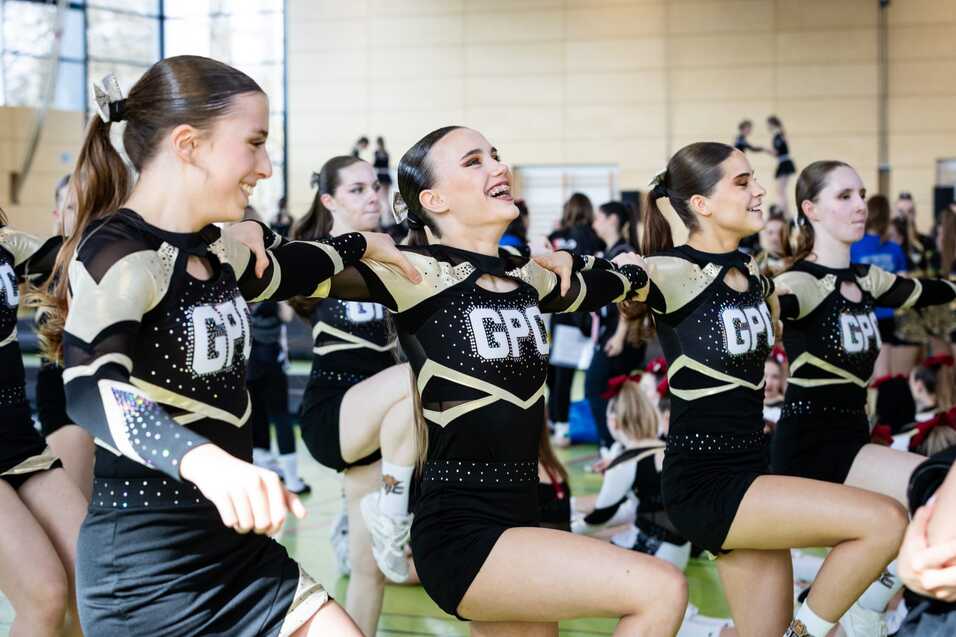 Vier Cheerleaderinnen in sportlicher Kleidung führen synchron eine Übung in einer Halle durch.